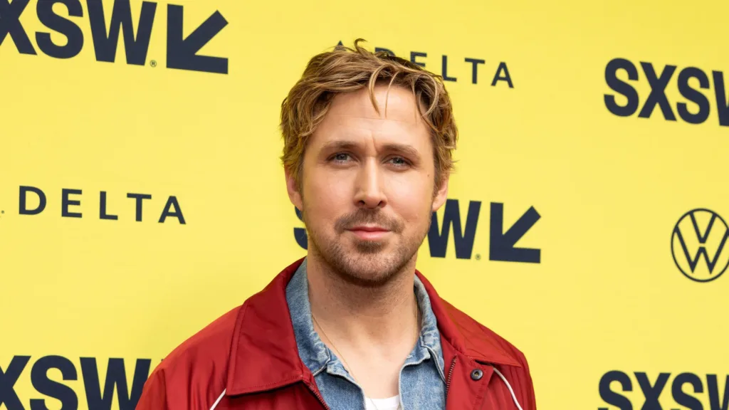 Ryan Gosling at the South by Southwest festival wearing blue denim shirt and red jacket. His jawline is no more sharp in the image as it was afew years back.